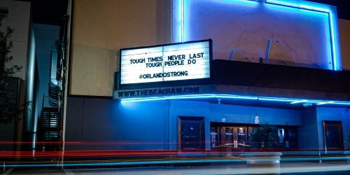 Tail lights streak past this night time photograph of a run down theatre, blue lights illuminate the doors, and a billboard above reads 'tough times don't last, tough people do' hashtag Orlando strong' How tough would they be if they new the ghosts under their feet.